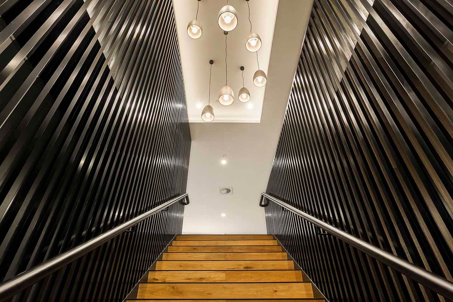 View of a modern staircase with wooden steps between two sleek, dark walls featuring horizontal slats. A series of pendant lights with round bulbs hang from the ceiling above, contributing to the contemporary design.