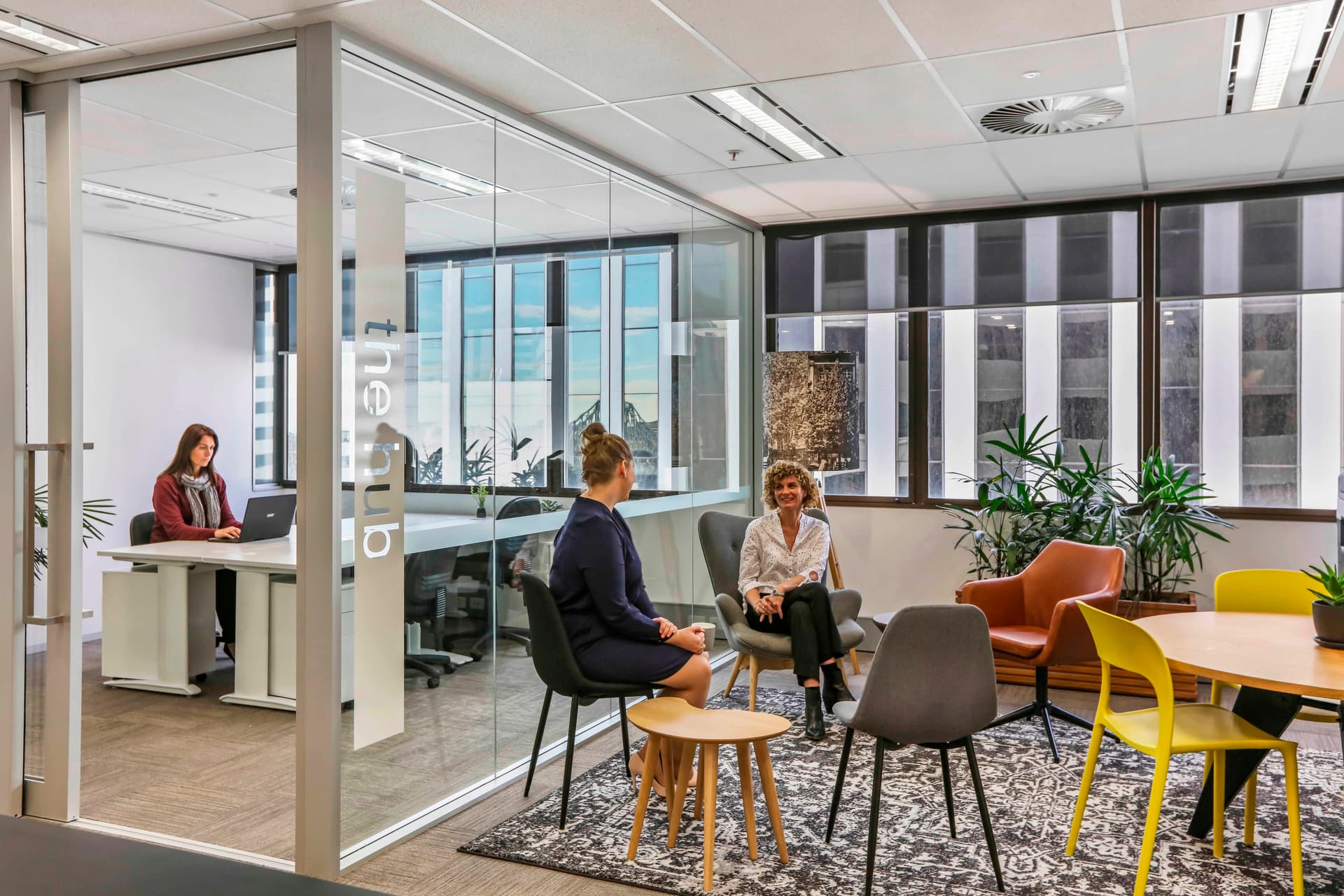 Modern office space with glass walls. Three people are conversing in a lounge area with colorful chairs and a rug. A fourth person works at a desk in an adjacent room labeled the club. Large windows provide natural light.