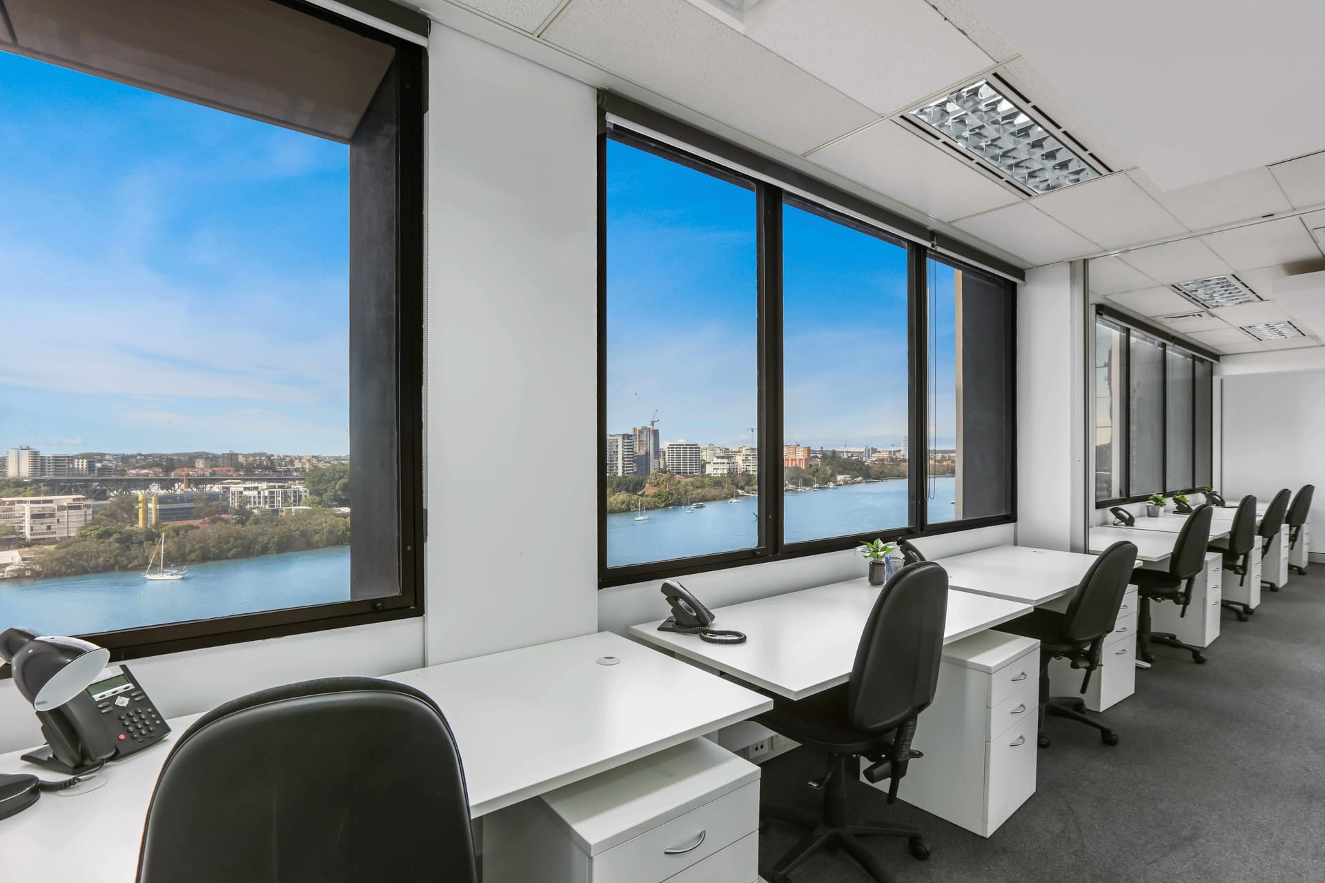 Office interior with rows of empty desks and chairs facing large windows. The windows offer a view of a river, cityscape, and clear blue sky. Phones and potted plants are on the desks, and the room has white walls and a gray carpet.