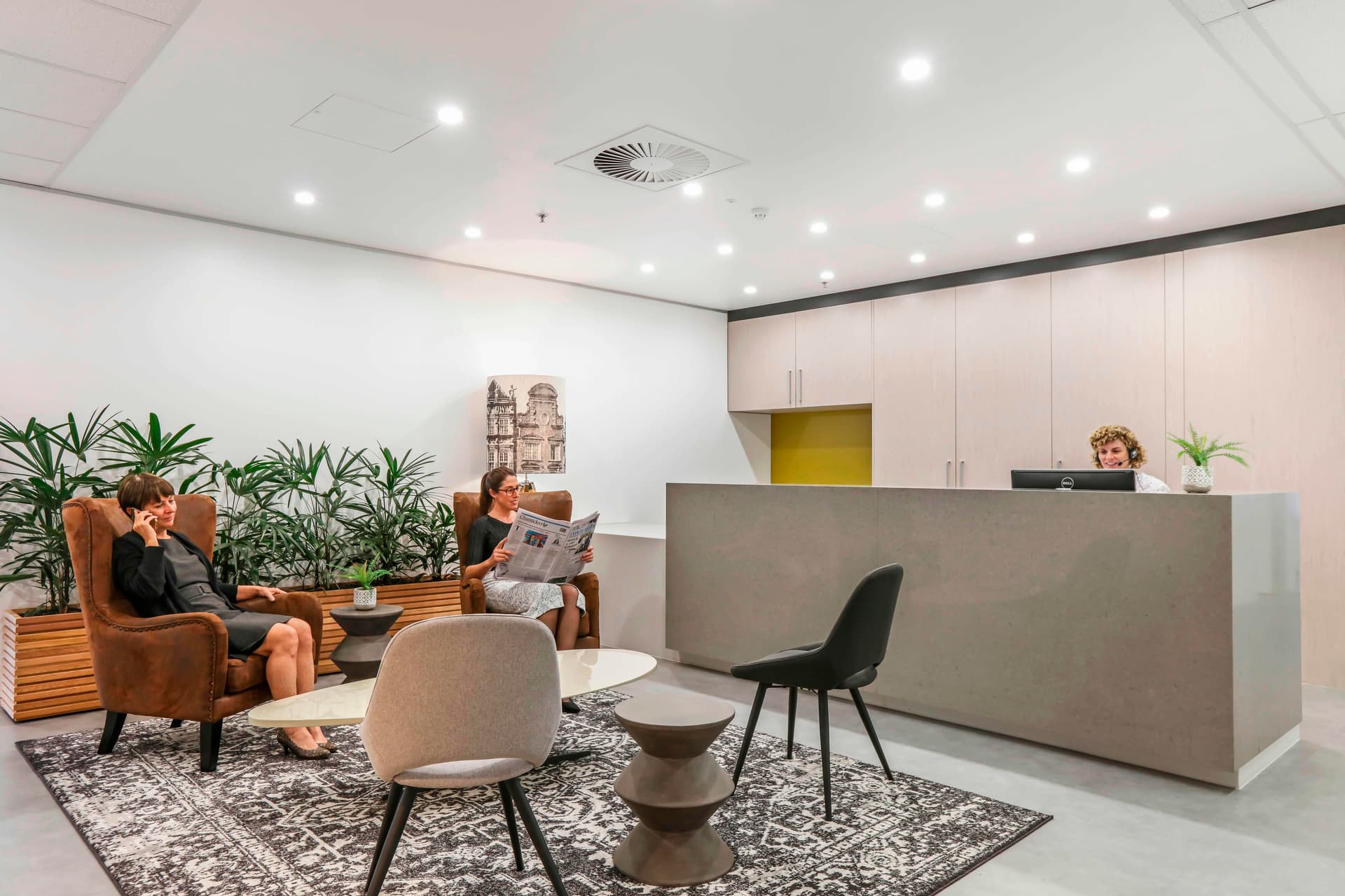 Modern waiting room with two women seated on leather chairs, reading. A reception desk is on the right, with a receptionist working. Potted plants decorate the area, and a patterned rug covers the floor. Bright, recessed lighting illuminates the space.