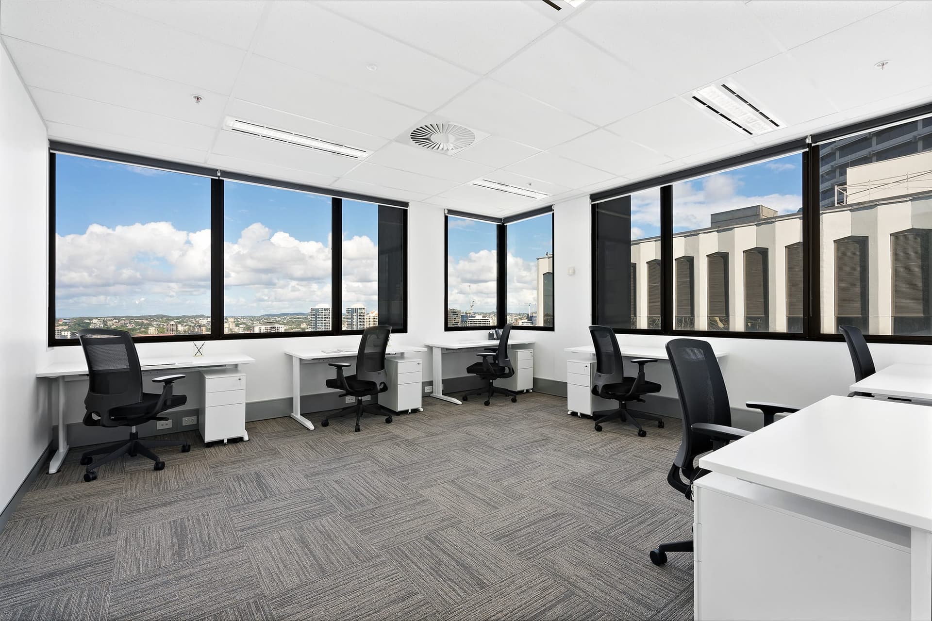 Modern office space with several white desks and black chairs, large windows offering a cityscape view. The room has a grey carpet and a clean, minimalist design with ample natural light.