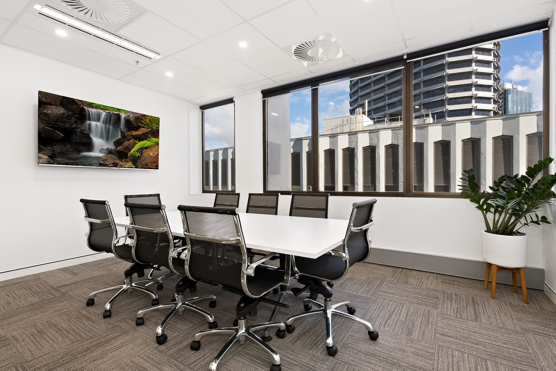 A modern conference room with a long white table and eight black office chairs. A large window showcases city buildings, and a television on the wall displays a nature scene. A green plant decorates the corner of the room.
