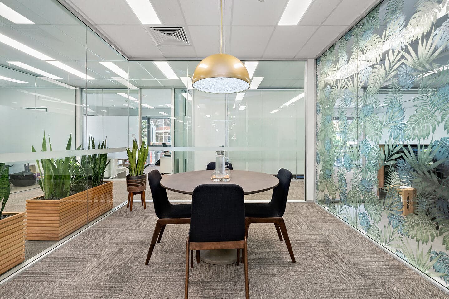 A modern office meeting room with a round table and four black chairs. A large gold pendant light hangs above the table. The room features glass walls with leafy designs and several potted plants. Neutral-colored carpet covers the floor.