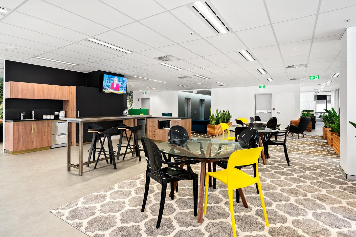Modern office break room with wooden cabinets, a countertop with stools, and tables with mixed chairs on patterned rugs. A wall-mounted TV is playing, and potted plants are arranged throughout the space.
