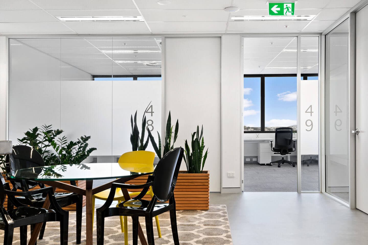 Modern office space with glass walls numbered 48 and 49, a table with yellow and black chairs, and potted plants. A window shows a blue sky outside. A green exit sign is visible above the doors.