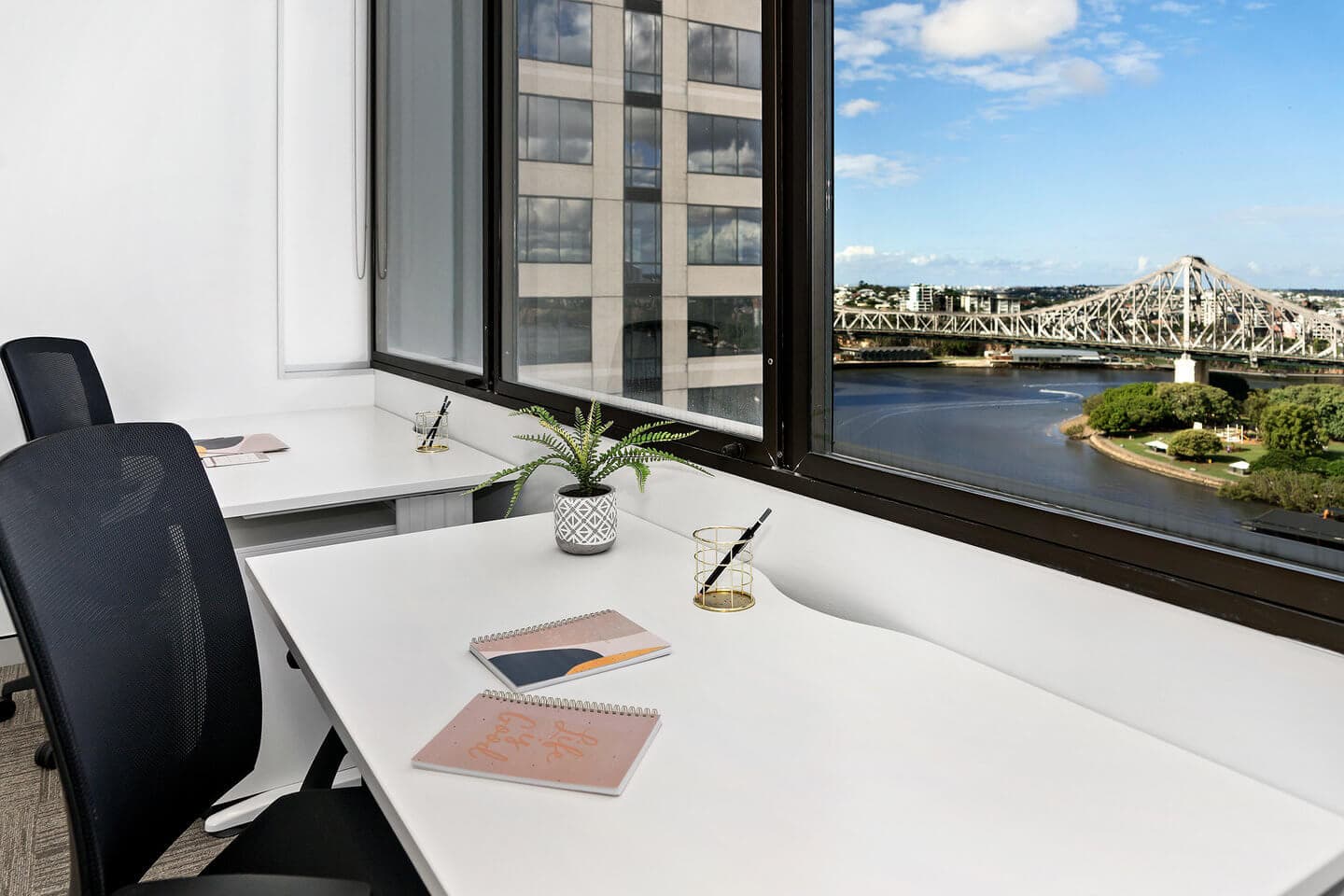 A modern office with two desks facing a large window, offering a view of a bridge over a river and nearby buildings. The desks have a small plant, notebooks, a pen holder, and a laptop. The sky is partly cloudy.