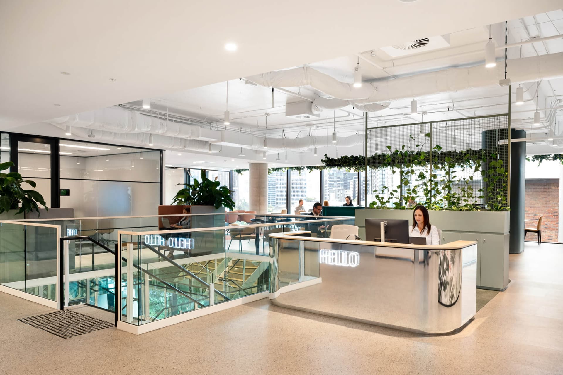 Modern office space with a glass-walled staircase, open seating, and vertical greenery. A person sits at a sleek reception desk with a HELLO sign. Large windows reveal a cityscape outside, and people work at tables in the background.