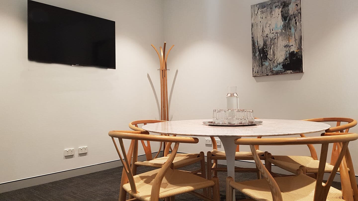 A minimalist meeting room with a round white table surrounded by four wooden chairs. A flat-screen TV is mounted on the wall above a coat rack. A large abstract painting adorns the adjacent wall. Natural lighting brightens the space.