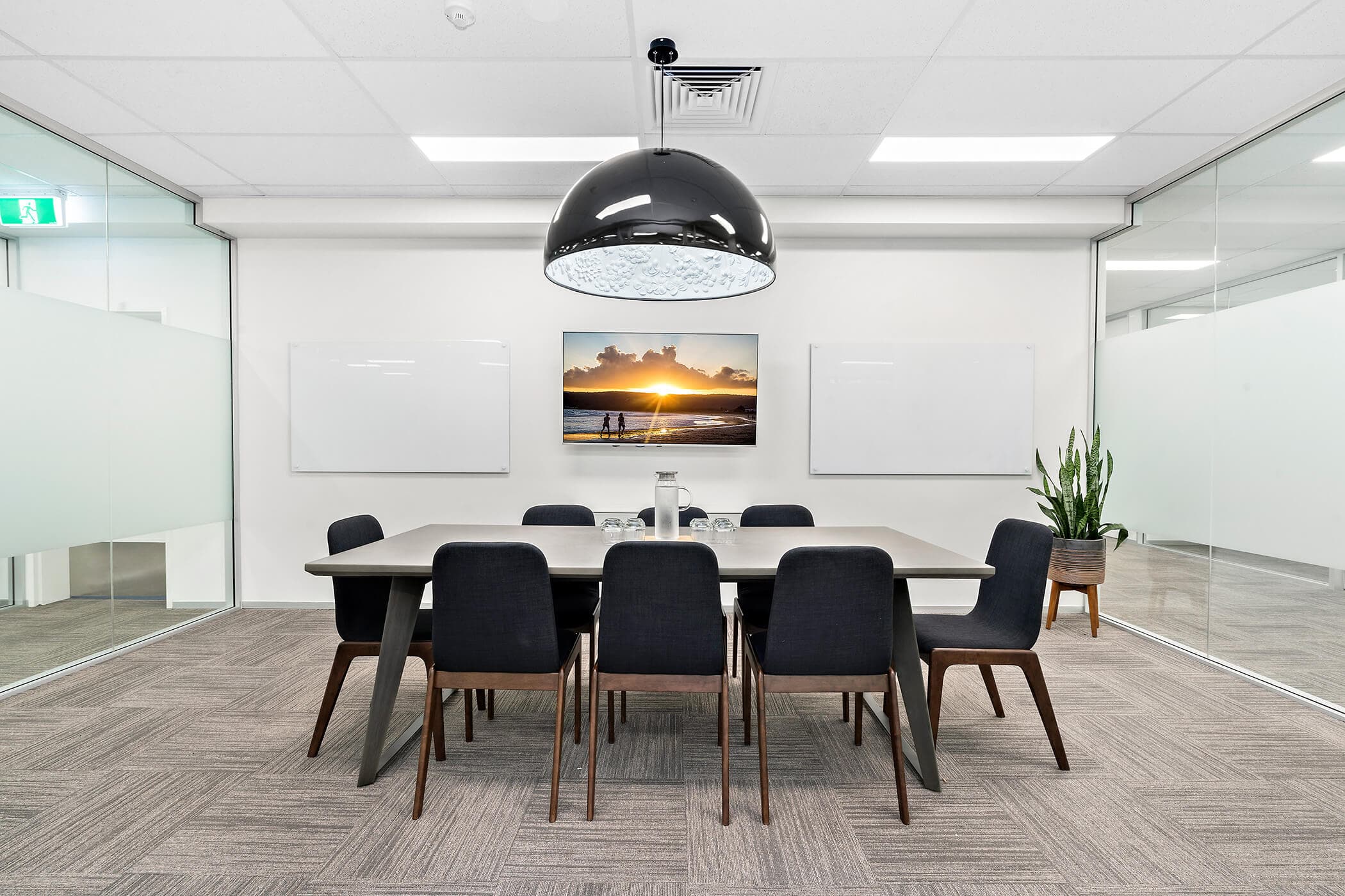 A modern conference room with a large table surrounded by eight dark chairs. A dome-shaped pendant light hangs above. Two blank whiteboards and a landscape photo adorn the walls. A potted plant is in the corner. Glass walls are on both sides.