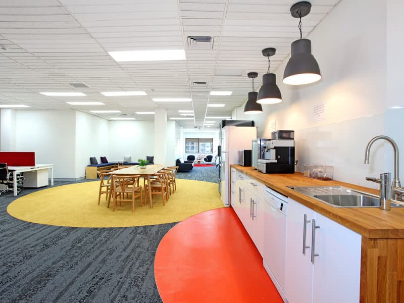 Modern office space with a kitchenette featuring a wooden countertop, coffee machine, and sink. A round table with wooden chairs sits on a yellow rug. Open workstations and lounge chairs are visible in the background, with ample natural light.