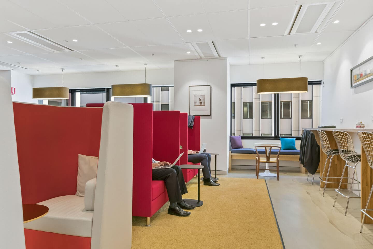 A modern office lounge with red semi-private booth seating, occupied by people working on laptops. Tables and chairs are arranged on a yellow carpet, with pendant lights hanging from the ceiling. Large windows let in natural light.