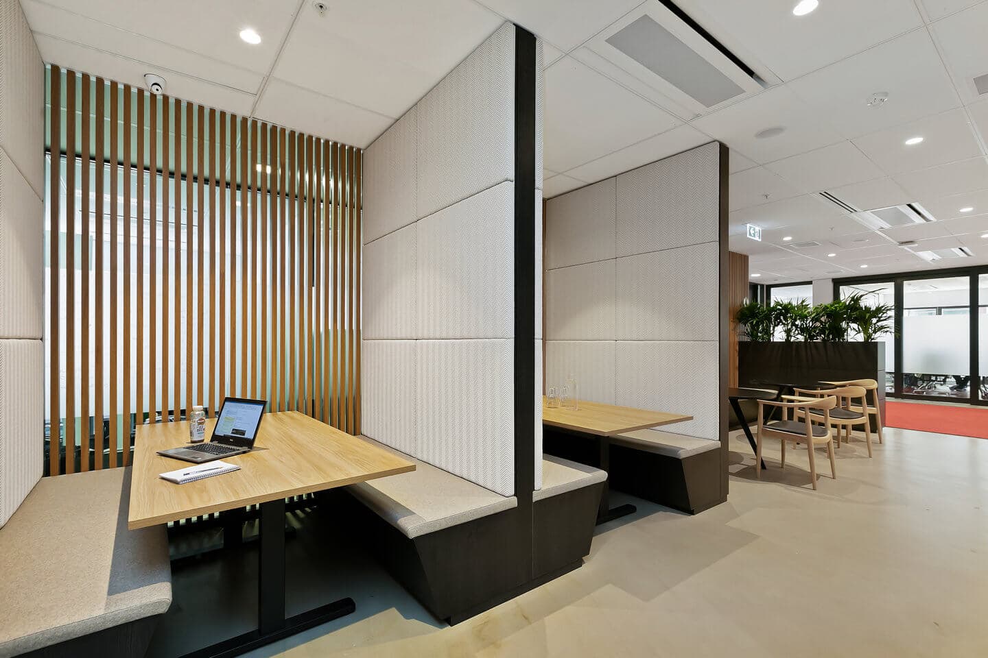 A modern office space with two booth seating areas featuring wooden tables and cushioned benches. A laptop and documents are on one table. Wooden slats and frosted glass windows enhance privacy. Indoor plants decorate the area in the background.
