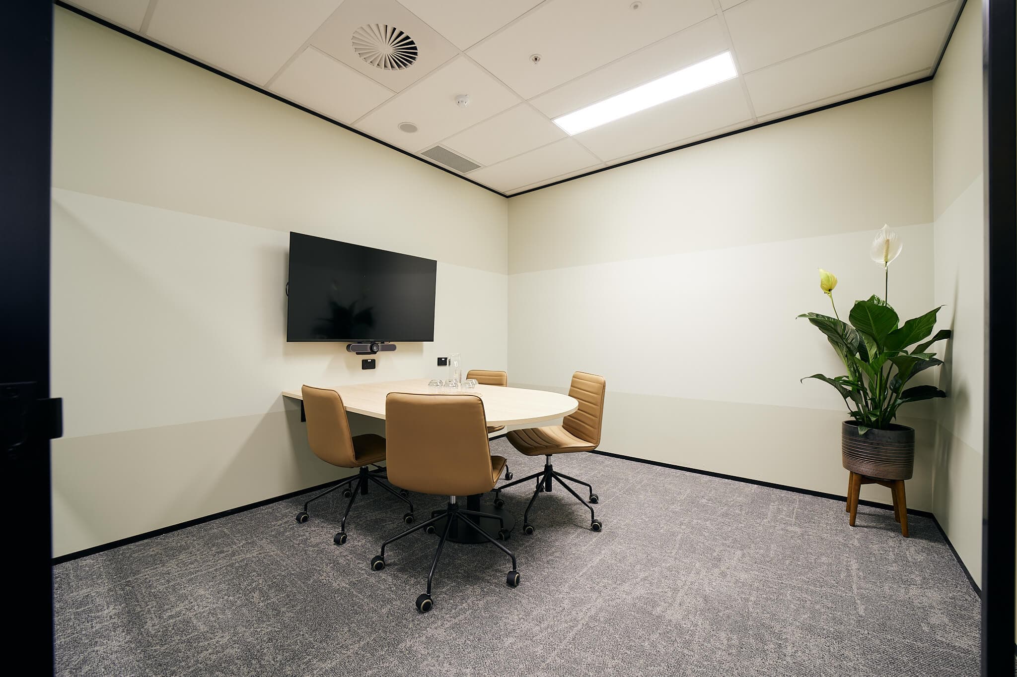 A small conference room with a round table and four beige chairs. A flat-screen TV is mounted on the wall. A potted plant is in the corner. The room has a carpeted floor and neutral-colored walls.