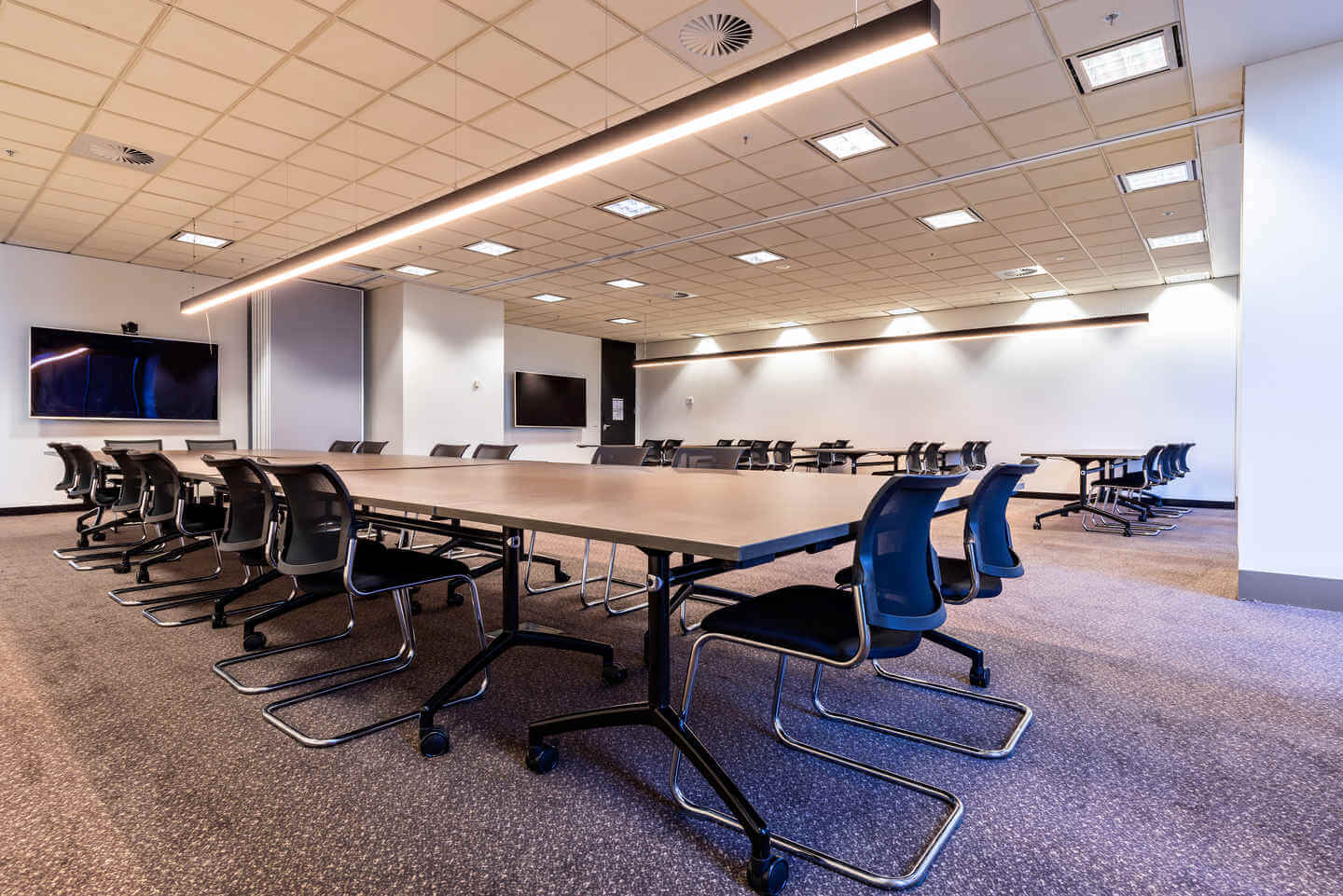 A modern conference room with long tables arranged in a U-shape, surrounded by black chairs. The room features two large screens on the walls and a contemporary overhead light fixture, with a beige carpeted floor and a tiled ceiling.