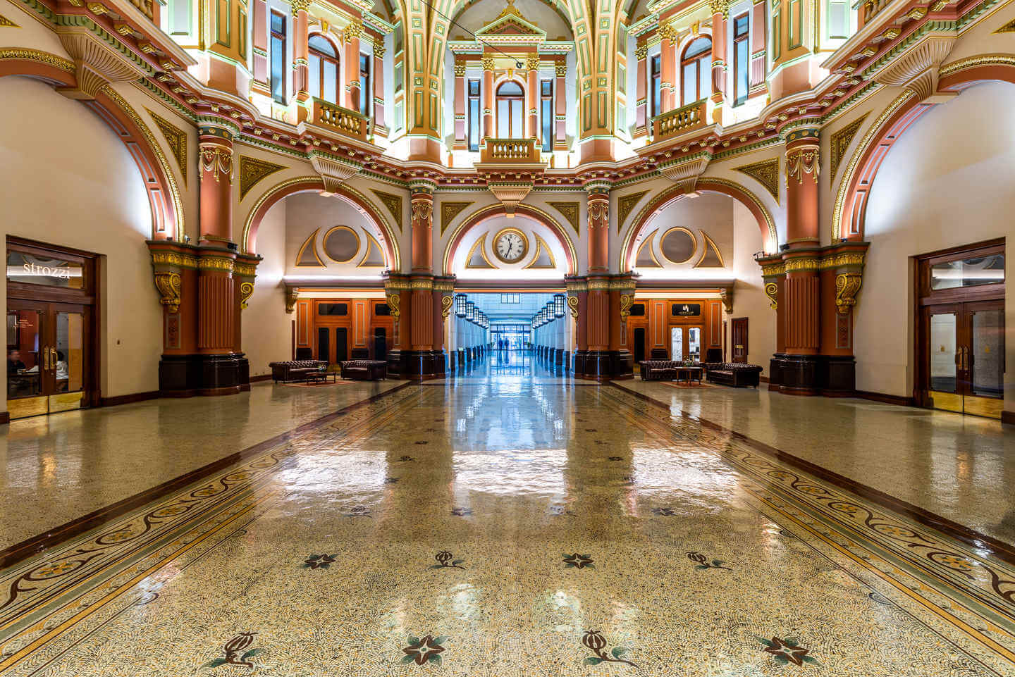 A grand interior hall with ornate architecture, large arches, and intricate mosaic floor. The ceiling features a detailed design with soft lighting. A staircase and balcony are visible in the background. The space is elegant and brightly lit.