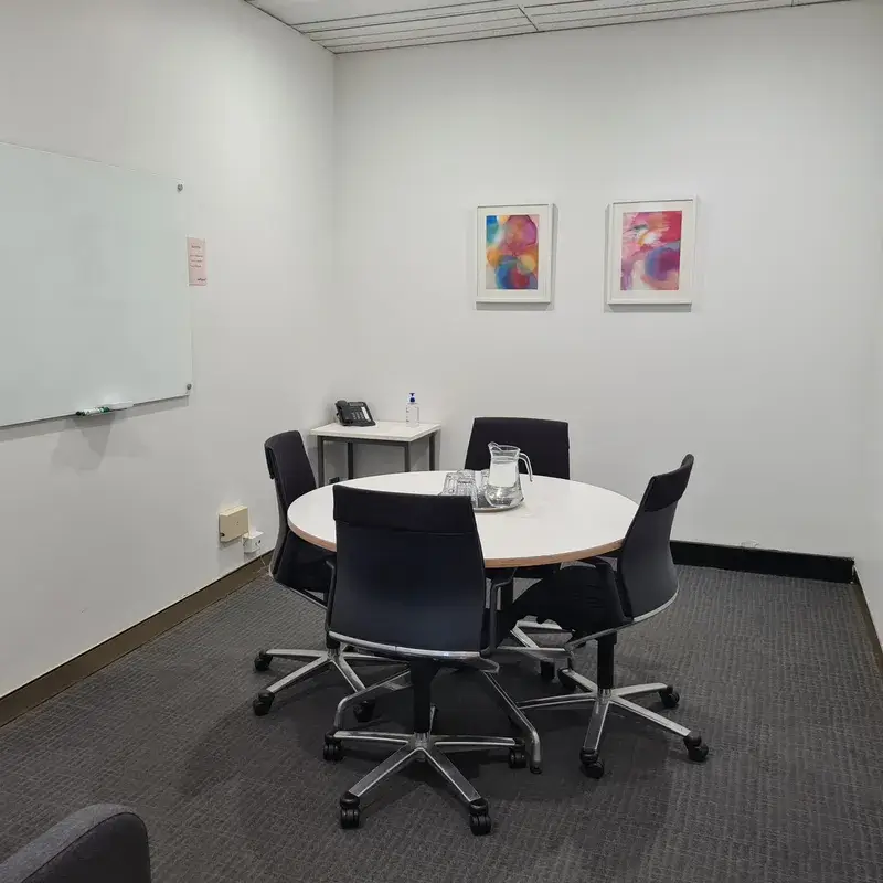 A small conference room with four chairs around a circular table. A pitcher with glasses is on the table. A whiteboard is mounted on the wall to the left. Two abstract art pieces hang on the white wall above a small corner table.