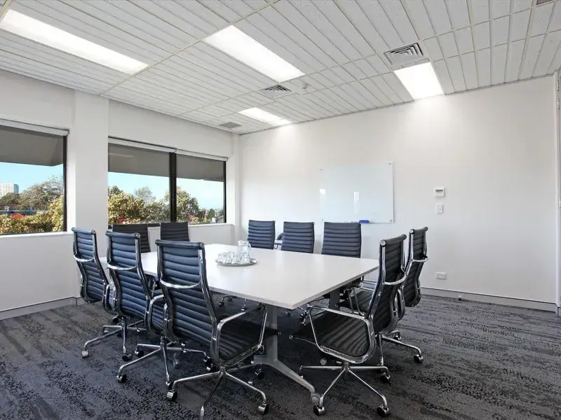 A modern conference room with a long rectangular table surrounded by ten black swivel chairs. Two large windows let in natural light. A whiteboard is mounted on one wall, and a glass pitcher with water and glasses sits on the table.
