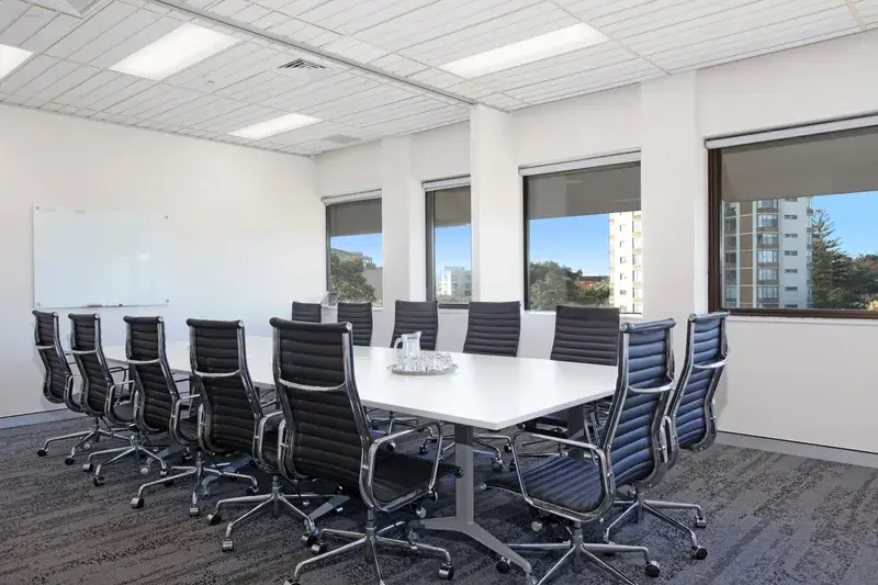 A modern conference room with a long white table surrounded by twelve black leather office chairs. There are large windows showing a view of nearby buildings, and a blank whiteboard on the wall. A tray with glasses sits on the table.