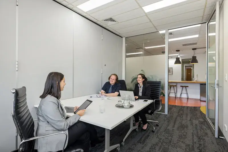 Three people sit around a white table in a modern office meeting room. Two people are on one side, facing the third person. There are laptops and notebooks on the table. Glass doors reveal a hallway and a counter with stools in the background.