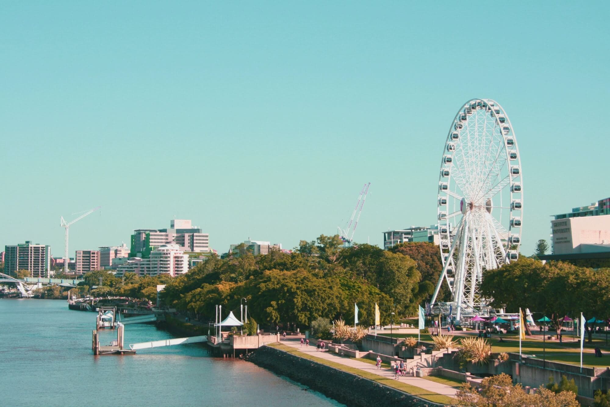 A scenic riverside view featuring a Ferris wheel, lush greenery, and modern buildings under a clear blue sky. The paths along the waters edge are lined with trees and people are strolling, enjoying the picturesque surroundings.
