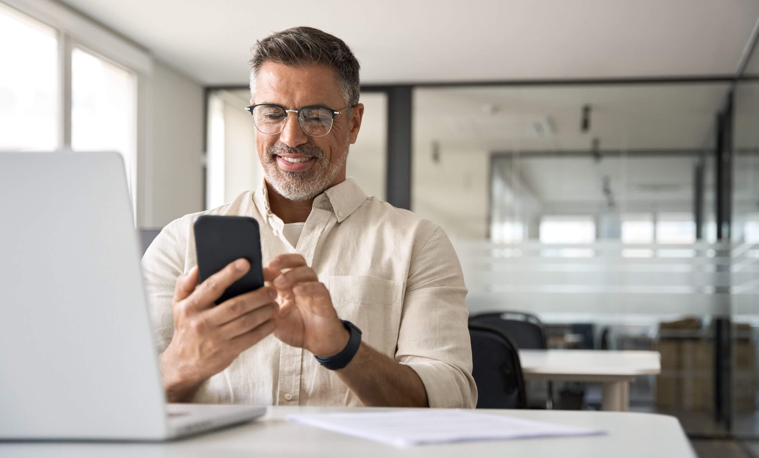 Middle aged Hispanic business manager ceo using cell phone mobile app, laptop. Smiling Latin or Indian mature man businessman holding smartphone sit in office working online on gadget with copy space.