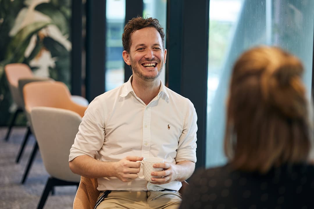 A man in a white shirt is sitting and smiling while holding a mug, engaged in conversation with a person whose back is to the camera. They are in a modern, well-lit room with plants and chairs in the background.