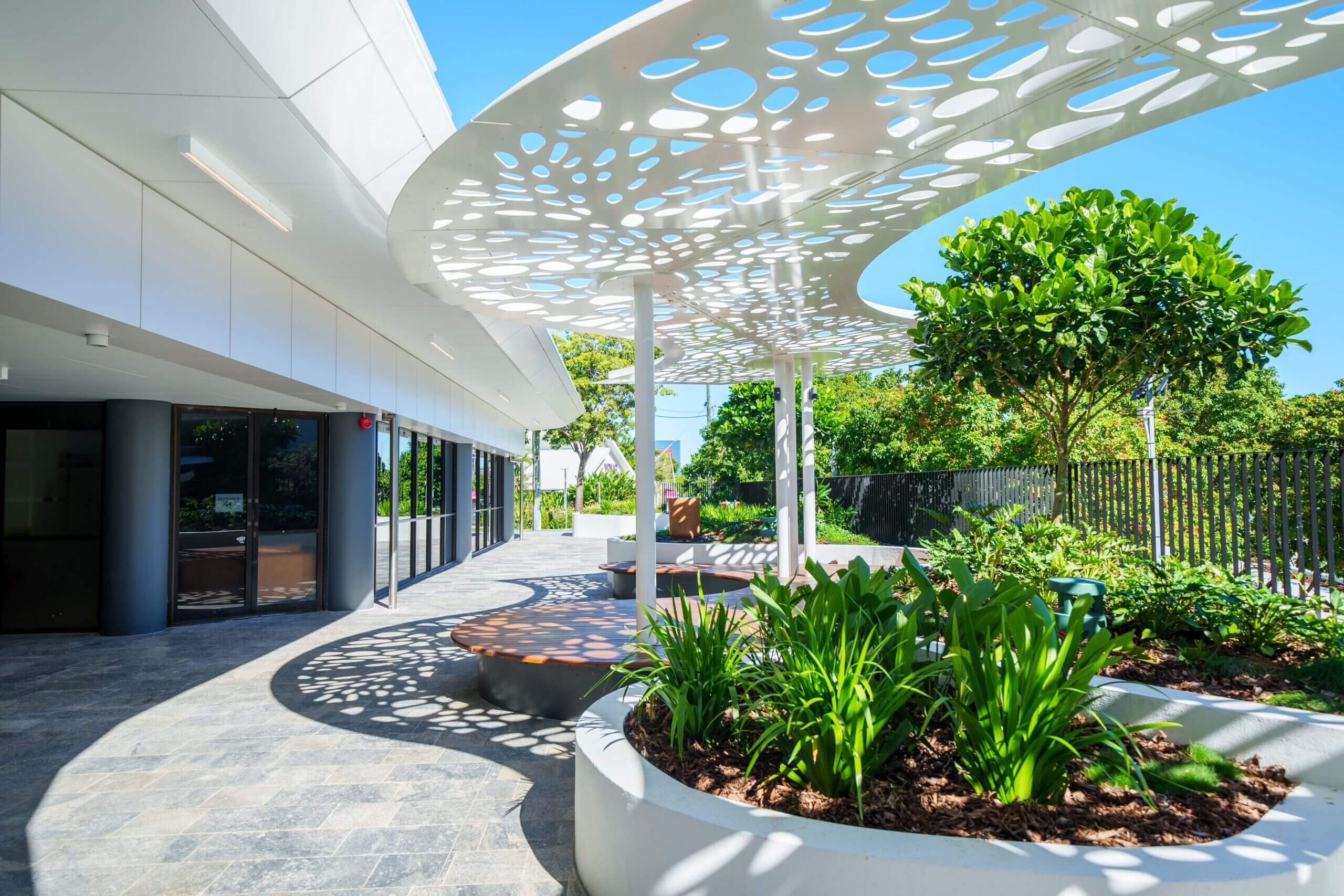 Modern outdoor space with perforated canopy casting patterned shadows on the ground. Includes curved planters filled with lush greenery, round benches, and a tiled stone floor. White building on the left, trees and a fence in the background.