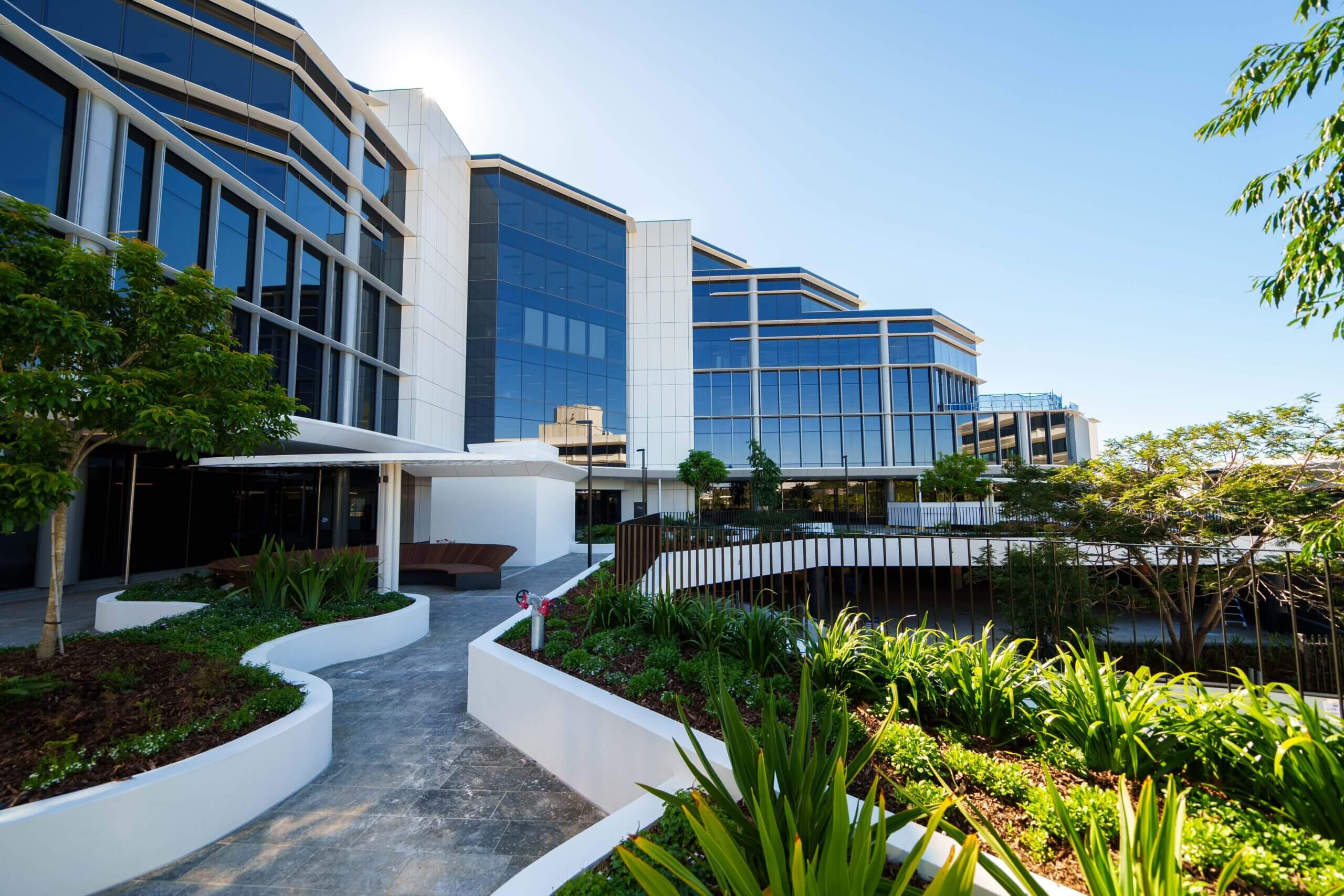Modern building exterior with reflective glass windows and geometric design. The foreground features landscaped gardens with green plants and winding pathways. Bright clear sky in the background.