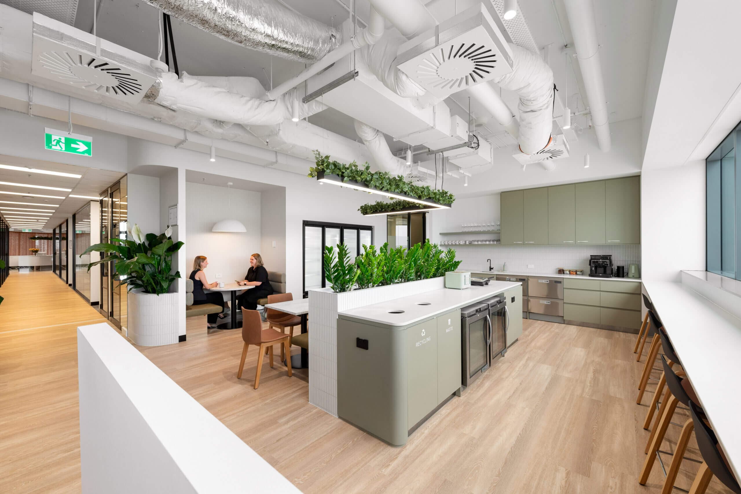 A brightly lit, contemporary office kitchen and lounge area. The design features an open ceiling with exposed ductwork painted white. The kitchen features a sage-green island with designated recycling and waste disposal areas, along with built-in refrigerators. Lush greenery adorns the top of the island. In the background, light sage-green cabinetry lines the wall, incorporating a sink and appliances. The far right of the image features a long counter bar with stools next to a window. To the left there is an area with two women sitting down at a table. An "Exit" sign directs from the doorway.