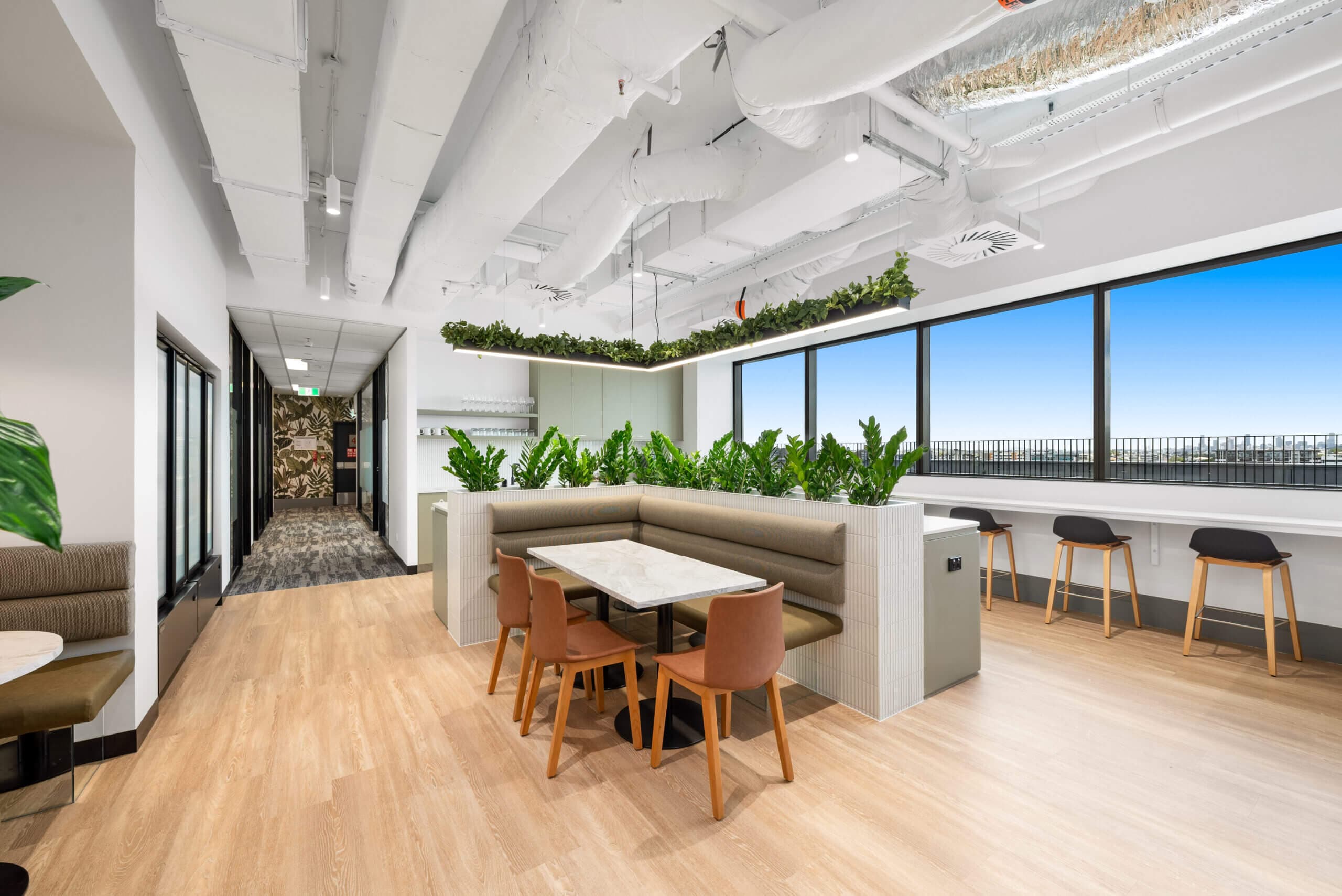 Modern office break area with booth seating, bar-height counter, and city view. Plants add greenery to the contemporary design.