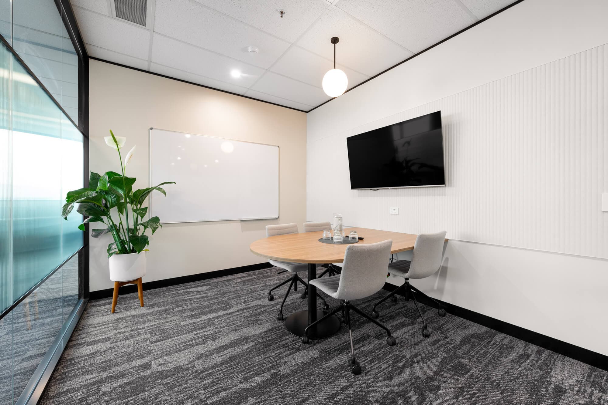 Modern conference room with round table, white board, TV, and plant. Neutral color scheme.