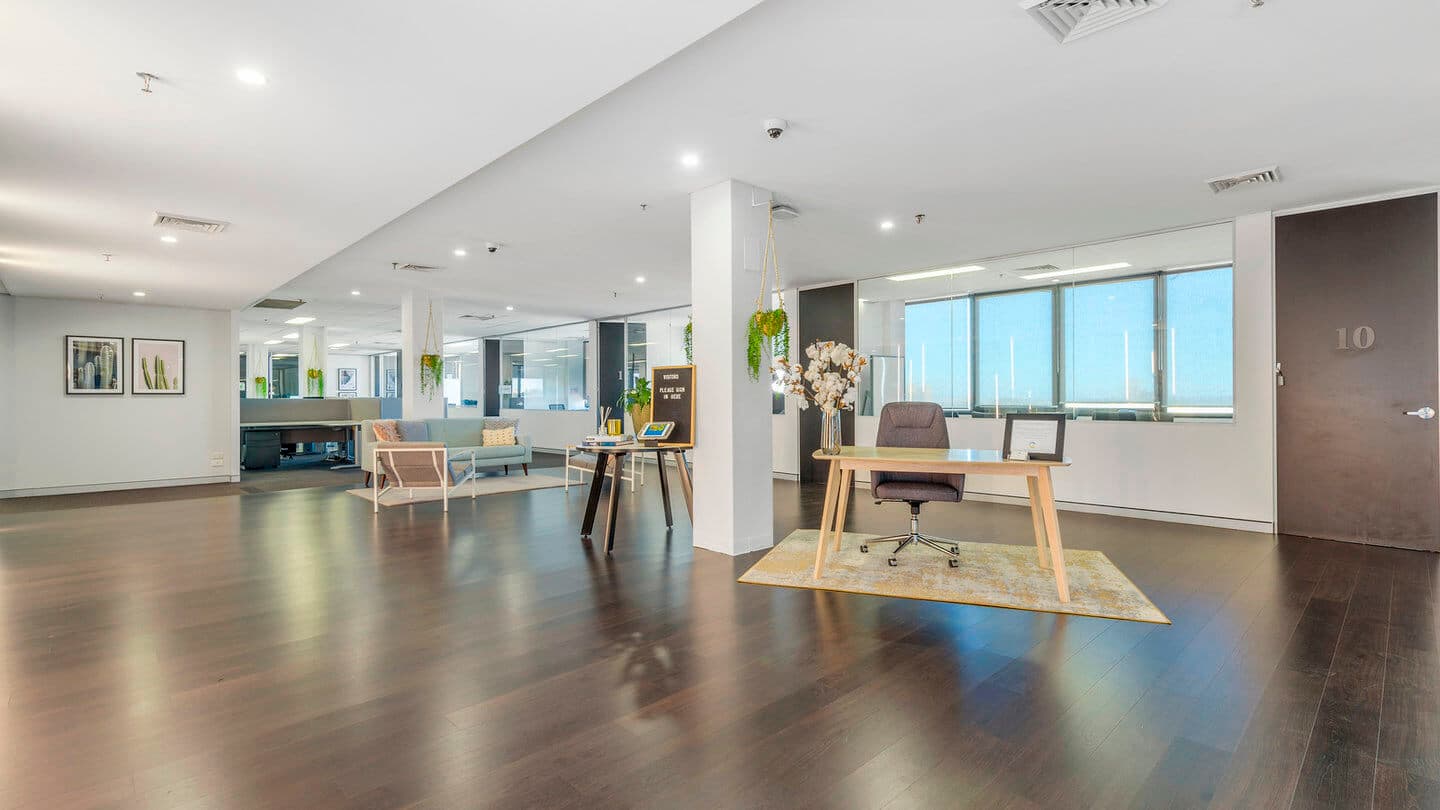 Spacious modern office with wooden floors, featuring a desk with a chair on a rug. Large windows offer bright, natural light. A seating area and decorations like plants and artwork add a welcoming touch.