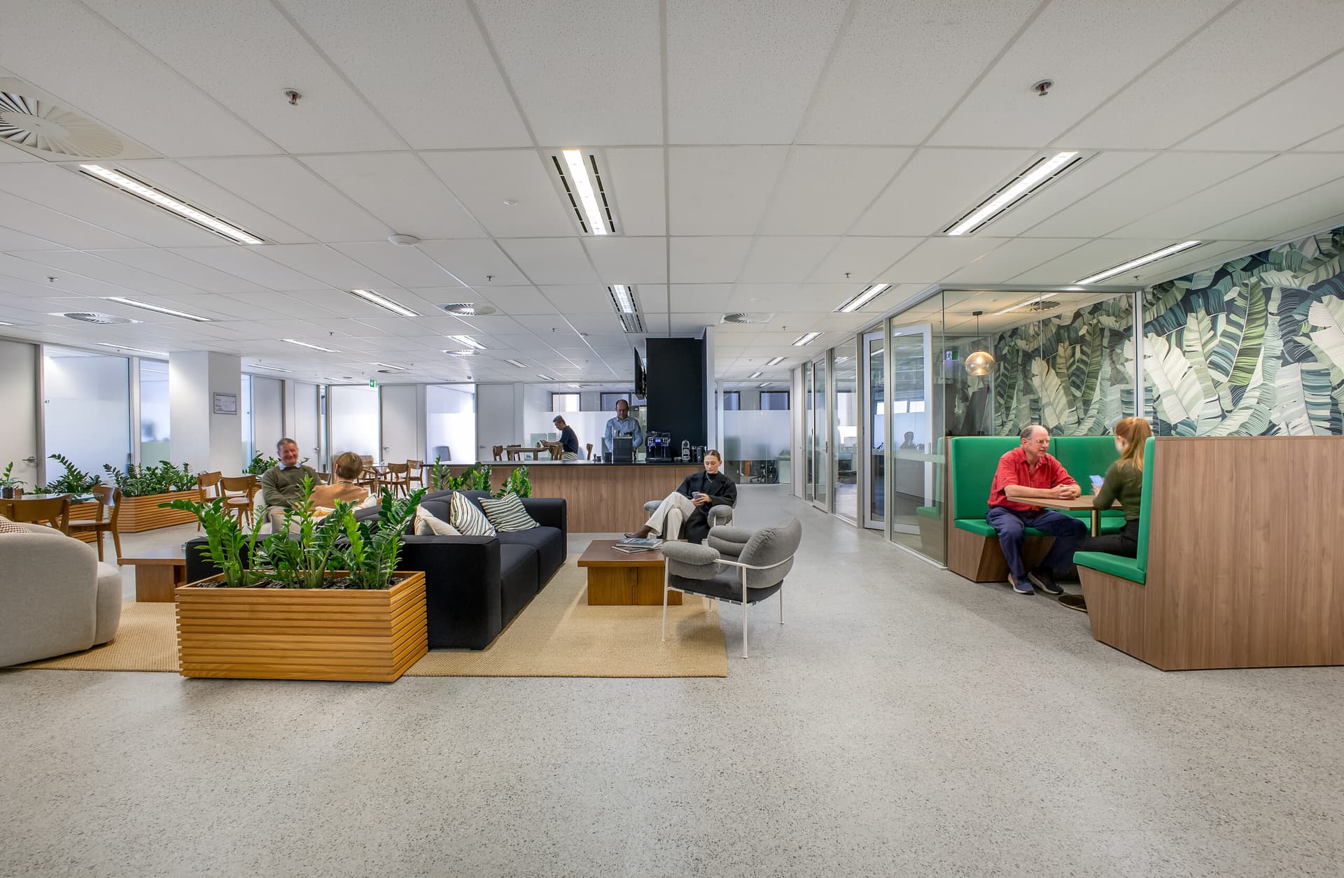 Modern open-plan office lounge with people sitting on couches, armchairs, and booth seats. Some are talking or working, with plants and large windows creating a bright, collaborative atmosphere.