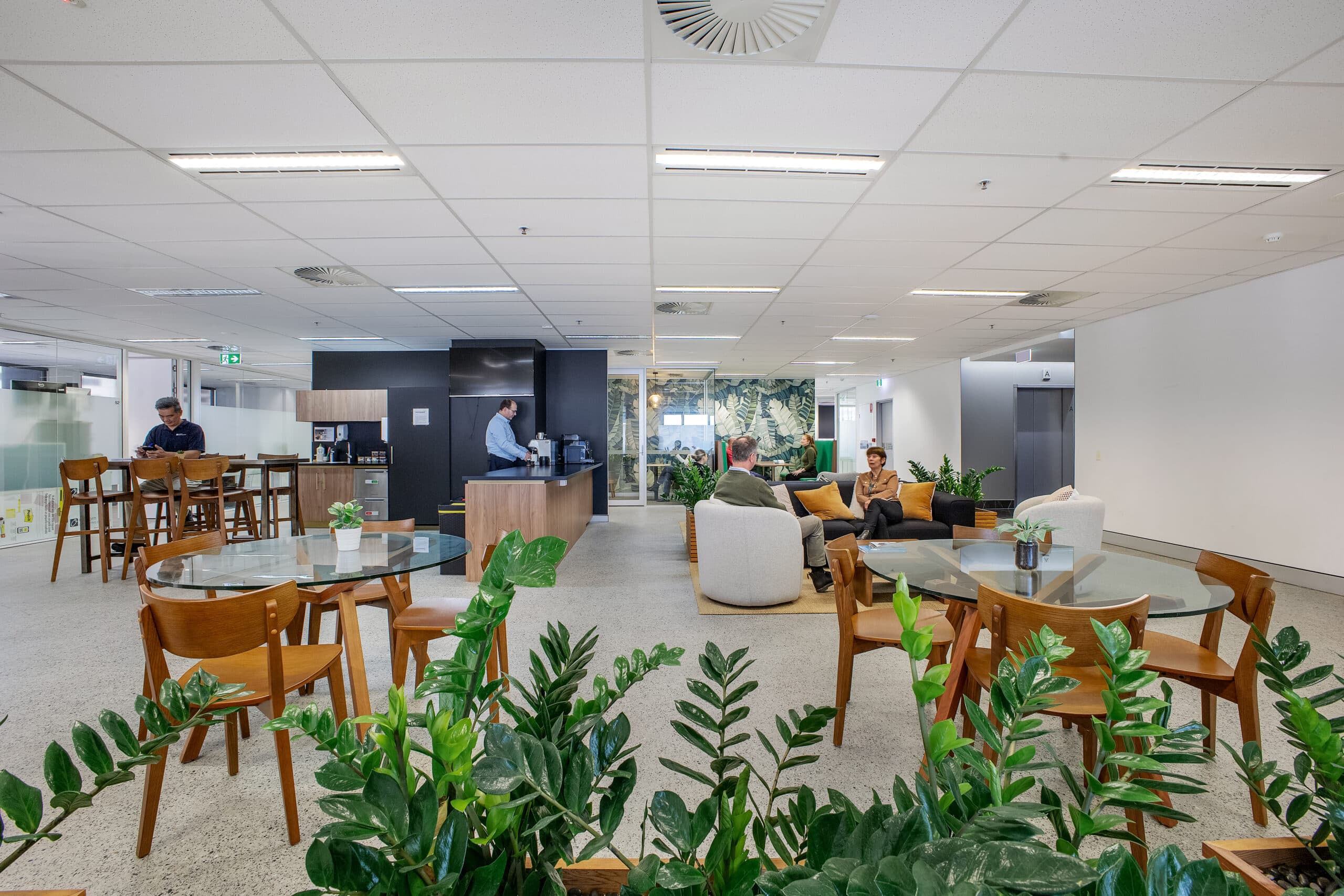 Modern office break room with several tables and chairs, green plants in the foreground, people sitting and talking, and a person at a counter in the background. Bright lighting and clean, open design.