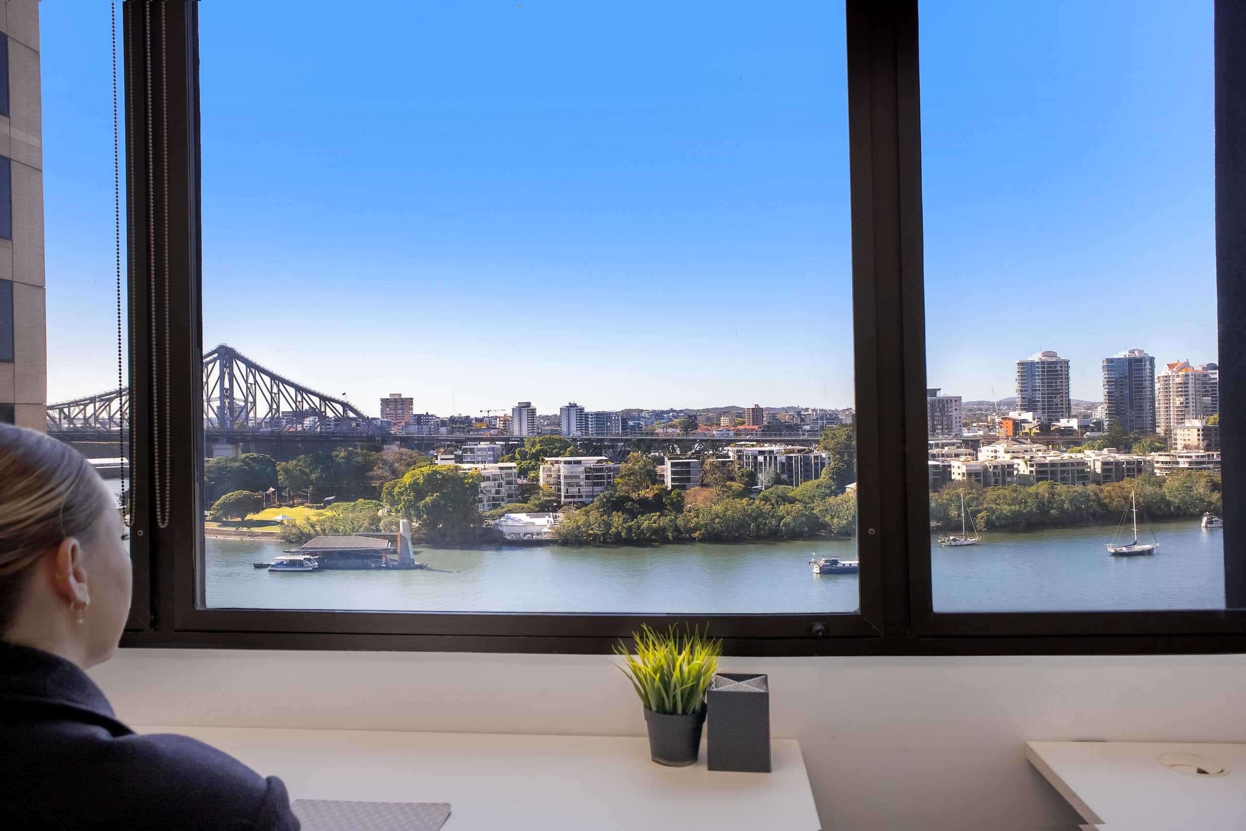 A person sits at a desk by a large window, looking out at a river, boats, and a cityscape with high-rise buildings and a bridge under a clear blue sky.