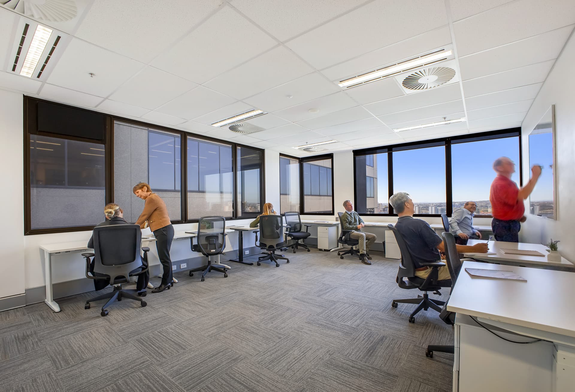 A modern office with six people working at desks and whiteboards. Some are seated and using laptops, while two stand and write on large whiteboards. Large windows let in natural light, and the office has a gray carpet and white walls.