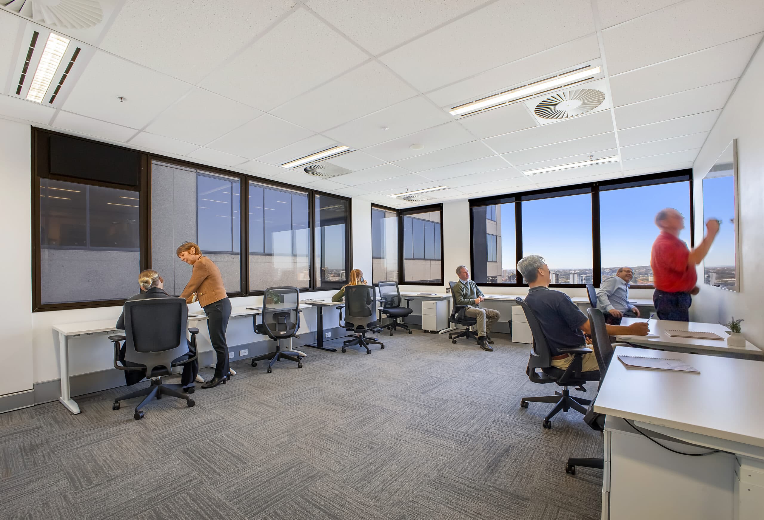A modern office with six people working at desks and whiteboards. Some are seated and using laptops, while two stand and write on large whiteboards. Large windows let in natural light, and the office has a gray carpet and white walls.