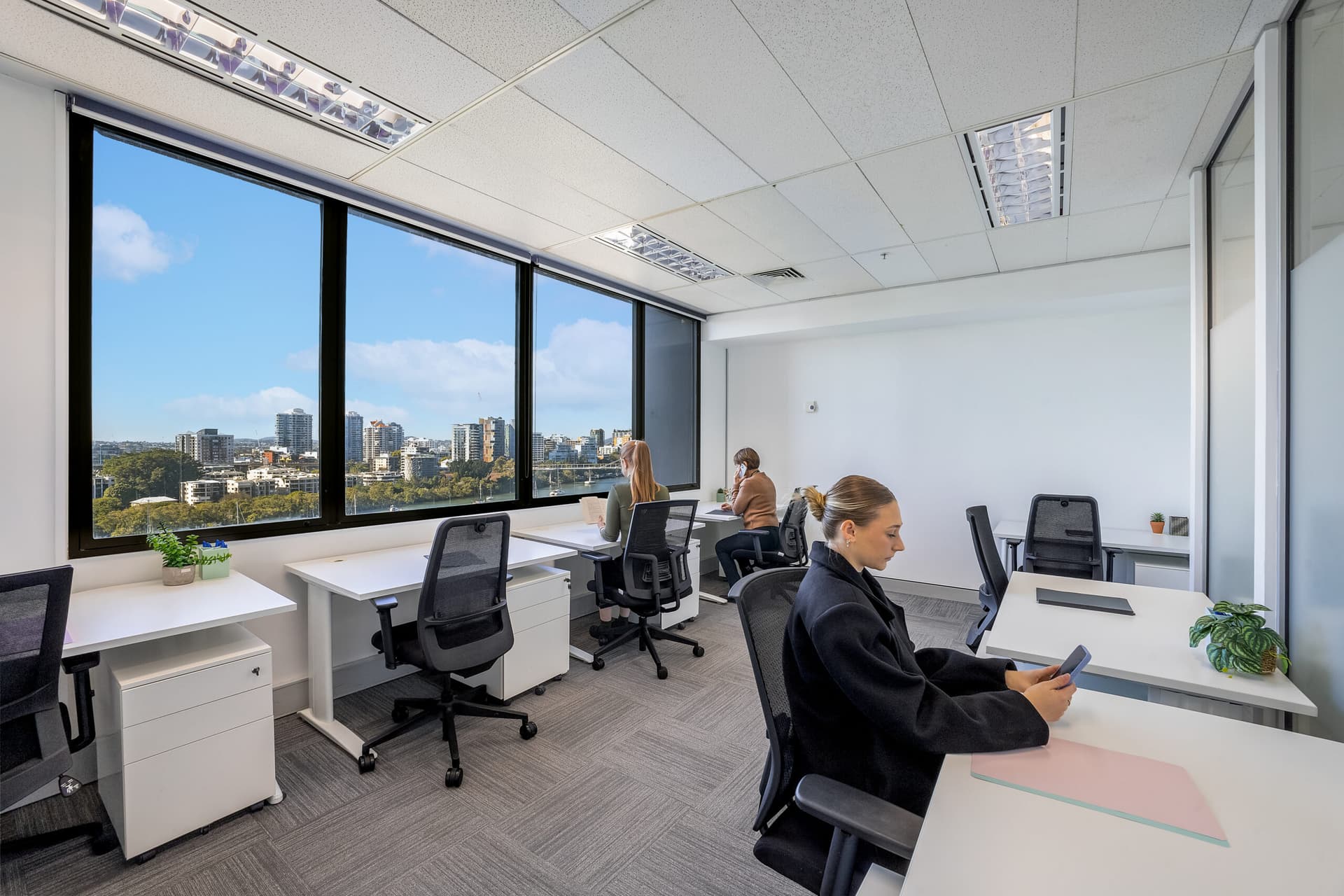 Modern office with large windows showing a city skyline. Four people are working at separate desks; one uses a phone, two are at computers, and one looks out the window. The room is bright with natural light and minimal decor.