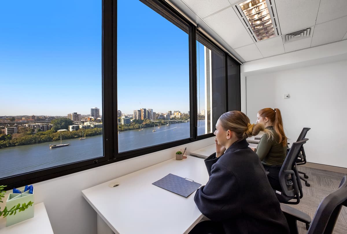 Two women sit at desks by large windows in a modern office, looking out at a city skyline and river with clear blue skies. The room is bright and minimalistic with simple office supplies on the desks.