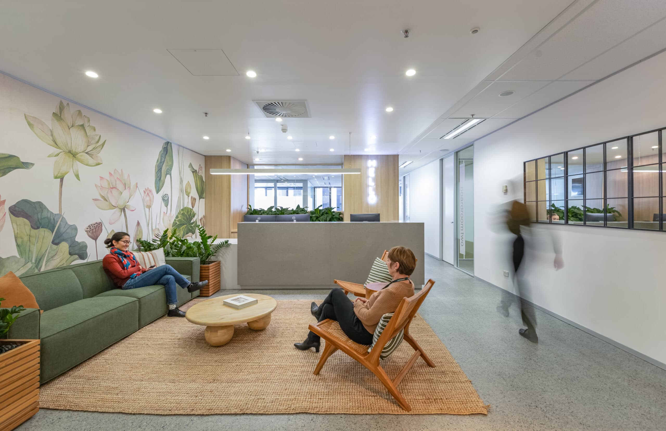 A modern office waiting area with two people sitting and talking, surrounded by plants and neutral furniture. A third person walks by. The space features a large floral wall mural and a reception desk in the background.