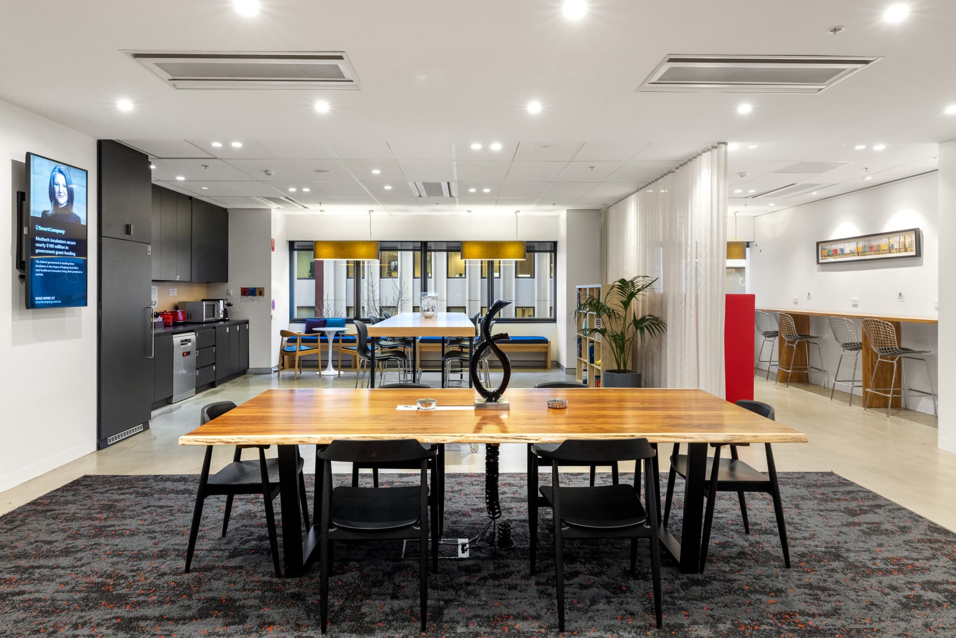 Modern office kitchen and dining area with a wooden table and black chairs in the foreground, a kitchenette on the left, a long bar with stools on the right, and large windows with yellow lamps in the background.