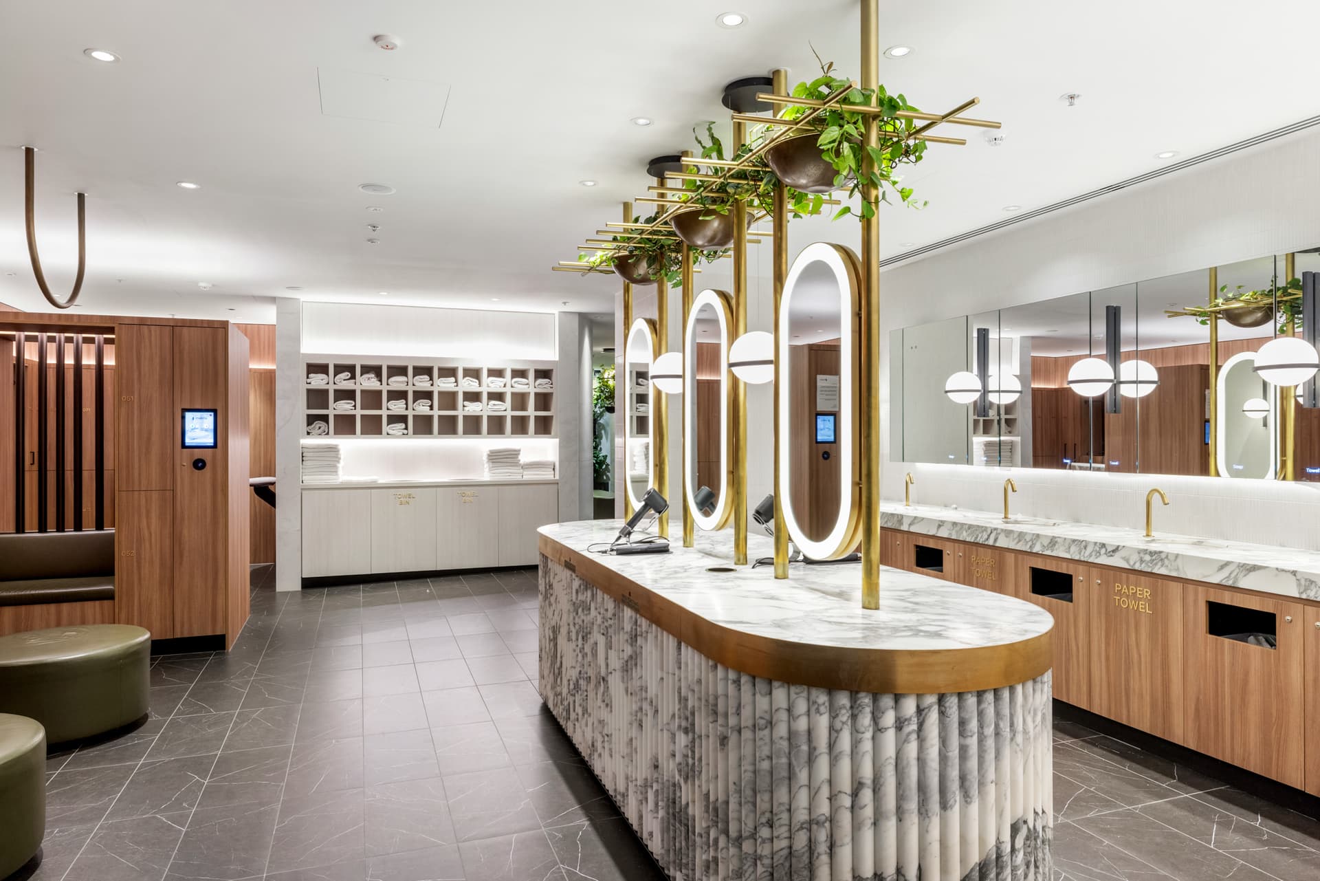 Modern restroom with marble countertops, oval mirrors, gold fixtures, and hanging plants. Wooden lockers and shelves line the walls, and the space is brightly lit with white and black tile flooring.