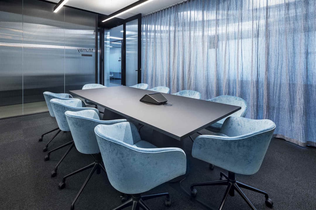 Modern conference room with a long black table surrounded by eight light blue upholstered chairs on wheels. A speakerphone is centered on the table. The room has glass walls and a sheer curtain, with soft overhead lighting.
