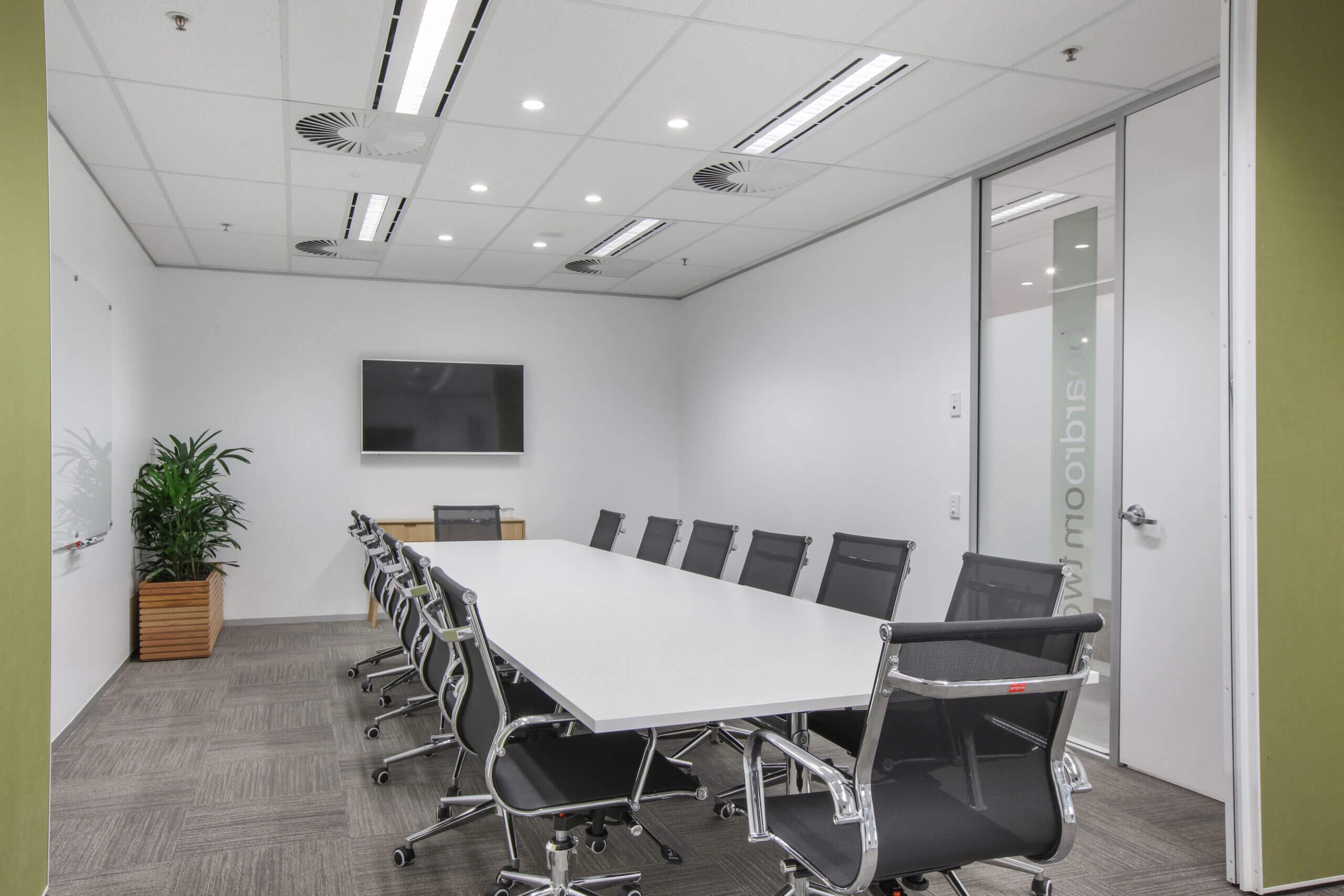 A modern conference room with a long white table and black office chairs. A flat-screen TV is mounted on the back wall, and a potted plant is in the corner. The walls are white, and the ceiling has recessed lighting.
