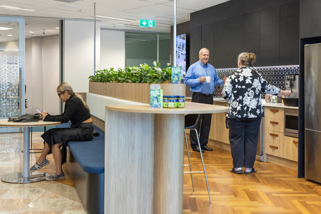 Three people are in a modern office kitchen. One woman sits on a bench using a tablet, while a man and another woman stand by the counter, talking and holding drinks. There are plants and kitchen appliances visible.