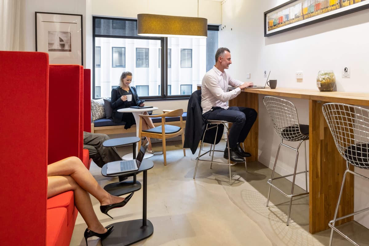 People working in a modern office space. A man sits at a high counter with a laptop, while others relax on lounge seating, engaging with laptops and coffee. The room has large windows, bright lighting, and an overall minimalist design.