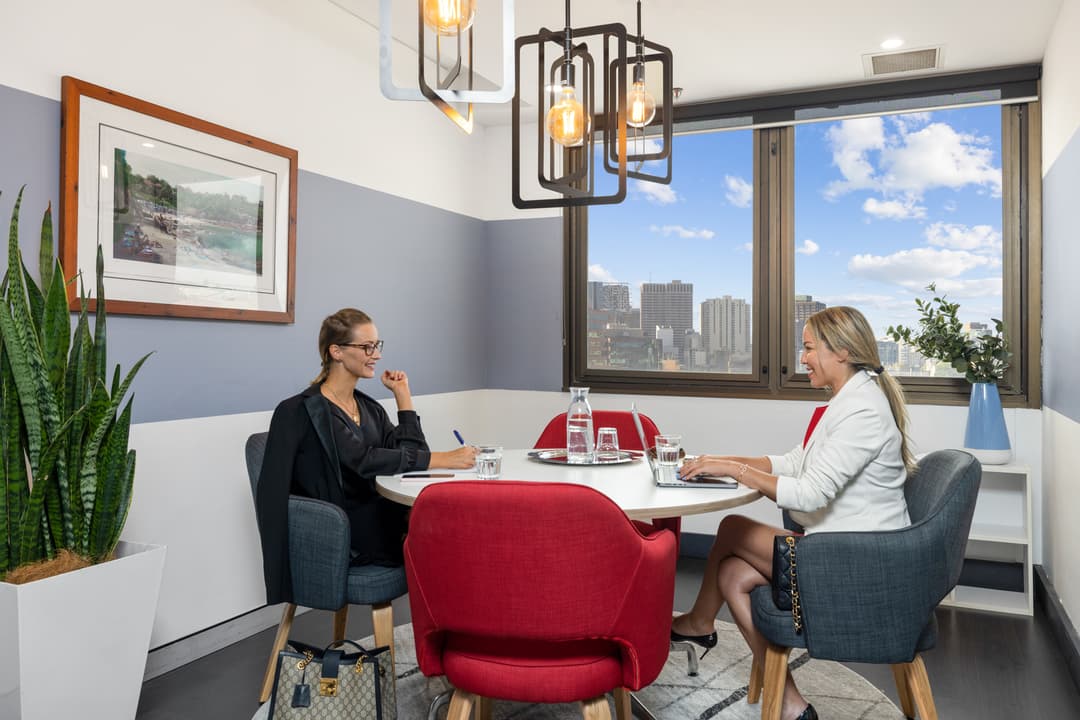 Two women in business attire sit across from each other at a round table in a modern office with large windows, red chairs, plants, notebooks, and water glasses. Cityscape visible outside on a sunny day.