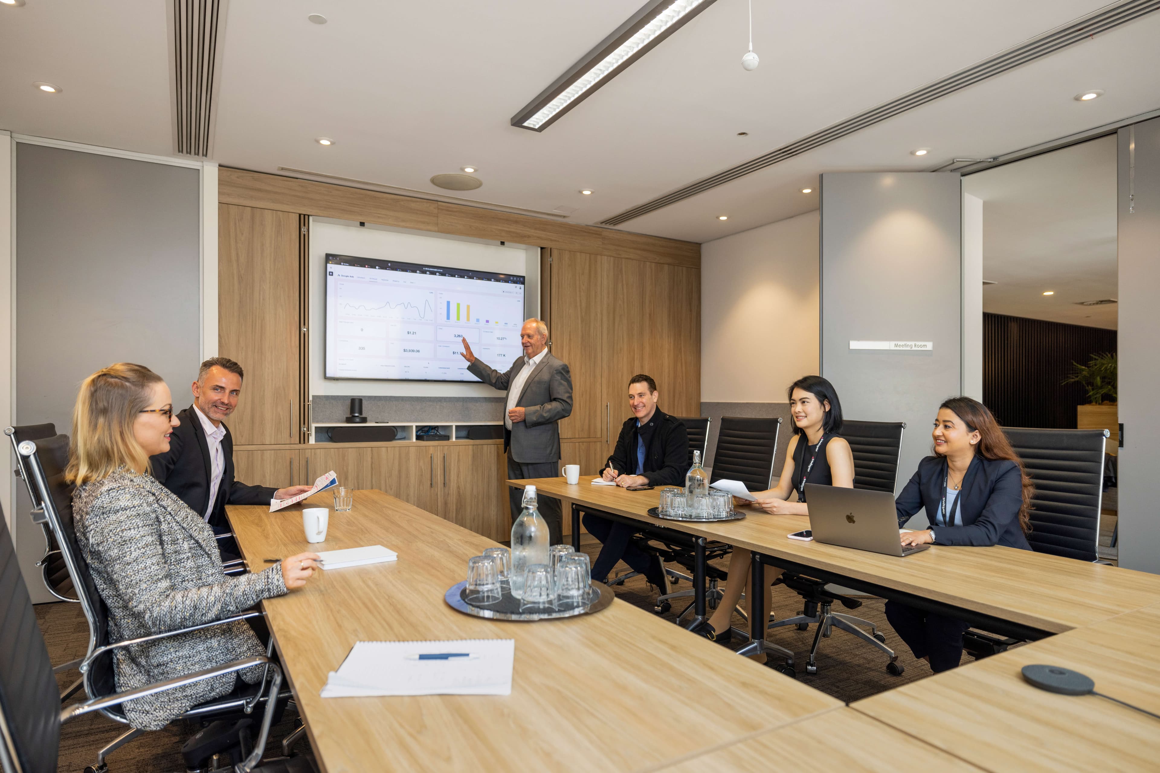 A modern conference room with a large wooden table surrounded by black chairs. A flat-screen TV displays an image of a bridge. The room features light wood paneling and has a bright atmosphere with overhead lighting.