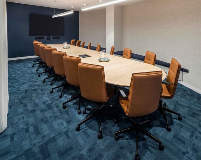 A modern conference room with a long oval table surrounded by brown leather chairs on a blue carpet. A large flat-screen TV is mounted on a dark accent wall at the end of the room. The room is well-lit with overhead lighting.