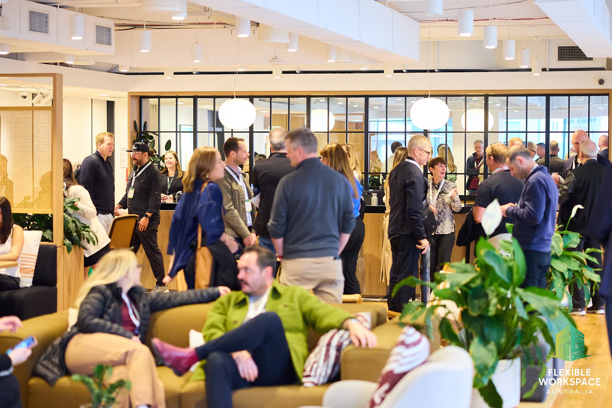 A group of people socialize and network in a modern, open office space with plants, lounge seating, and a reception counter. Some are seated, while others stand in conversation. The Flexible Workspace Australia logo appears in the corner.